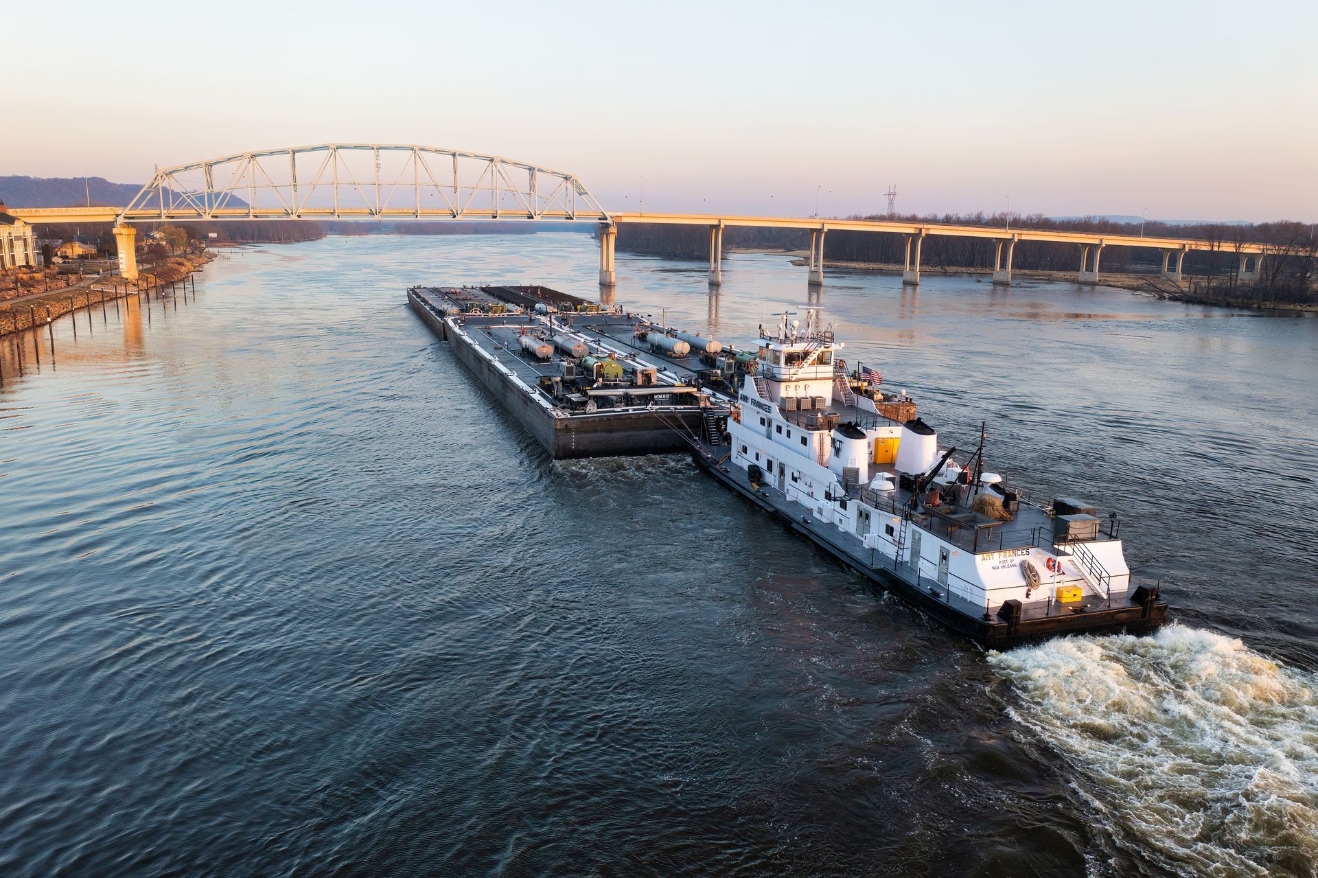 Tugboat pushing a barge along a river at sunset, with a bridge in the background.