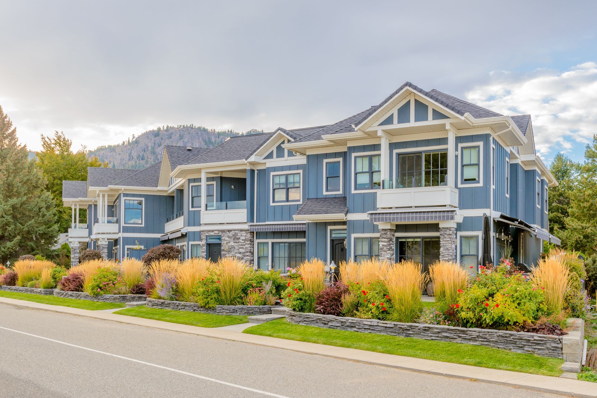 Modern blue apartment building with landscaped shrubs along a street, backed by mountains