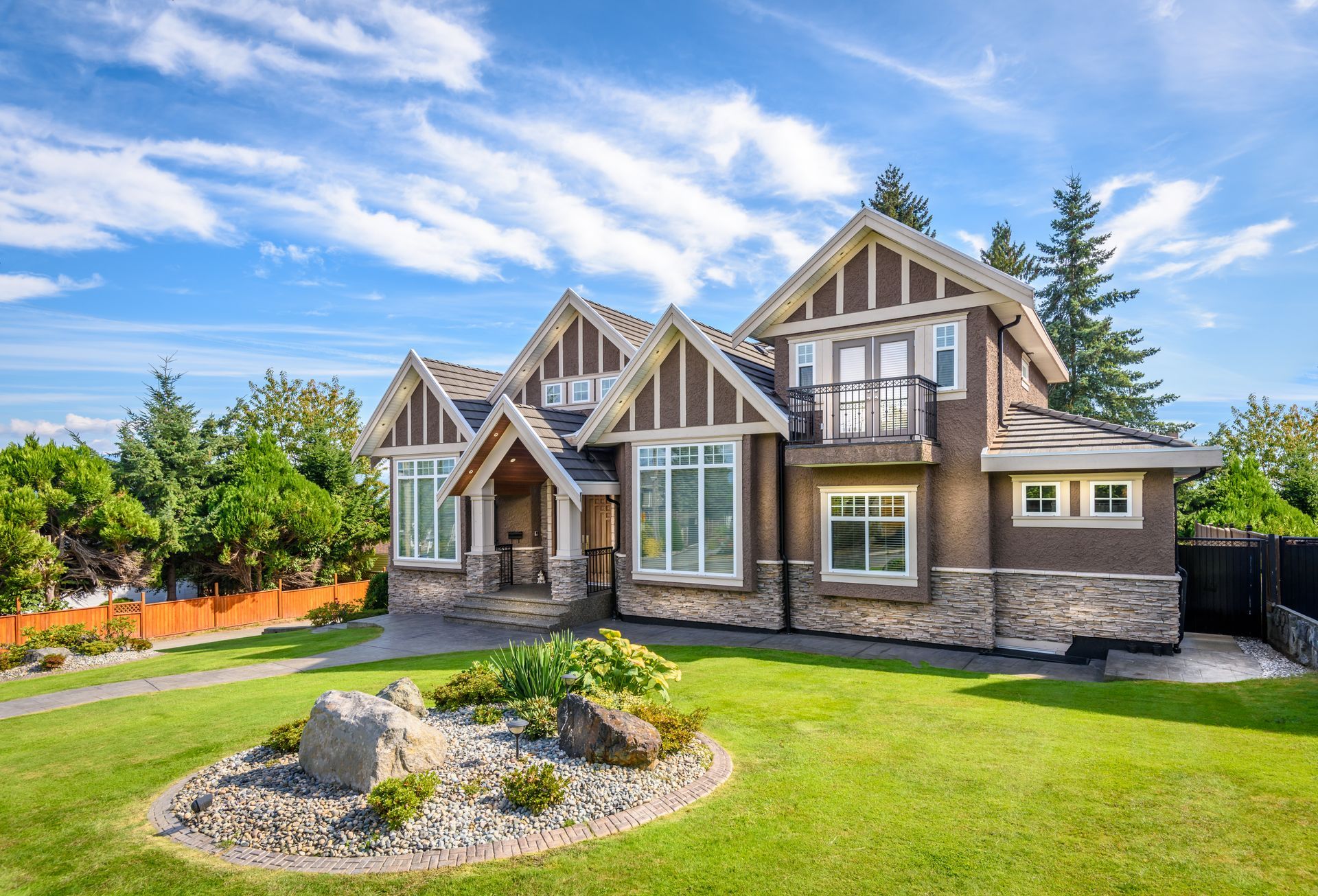 Modern Craftsman-style house with stone and wood exterior, manicured lawn, and landscaped front yard under a blue sky