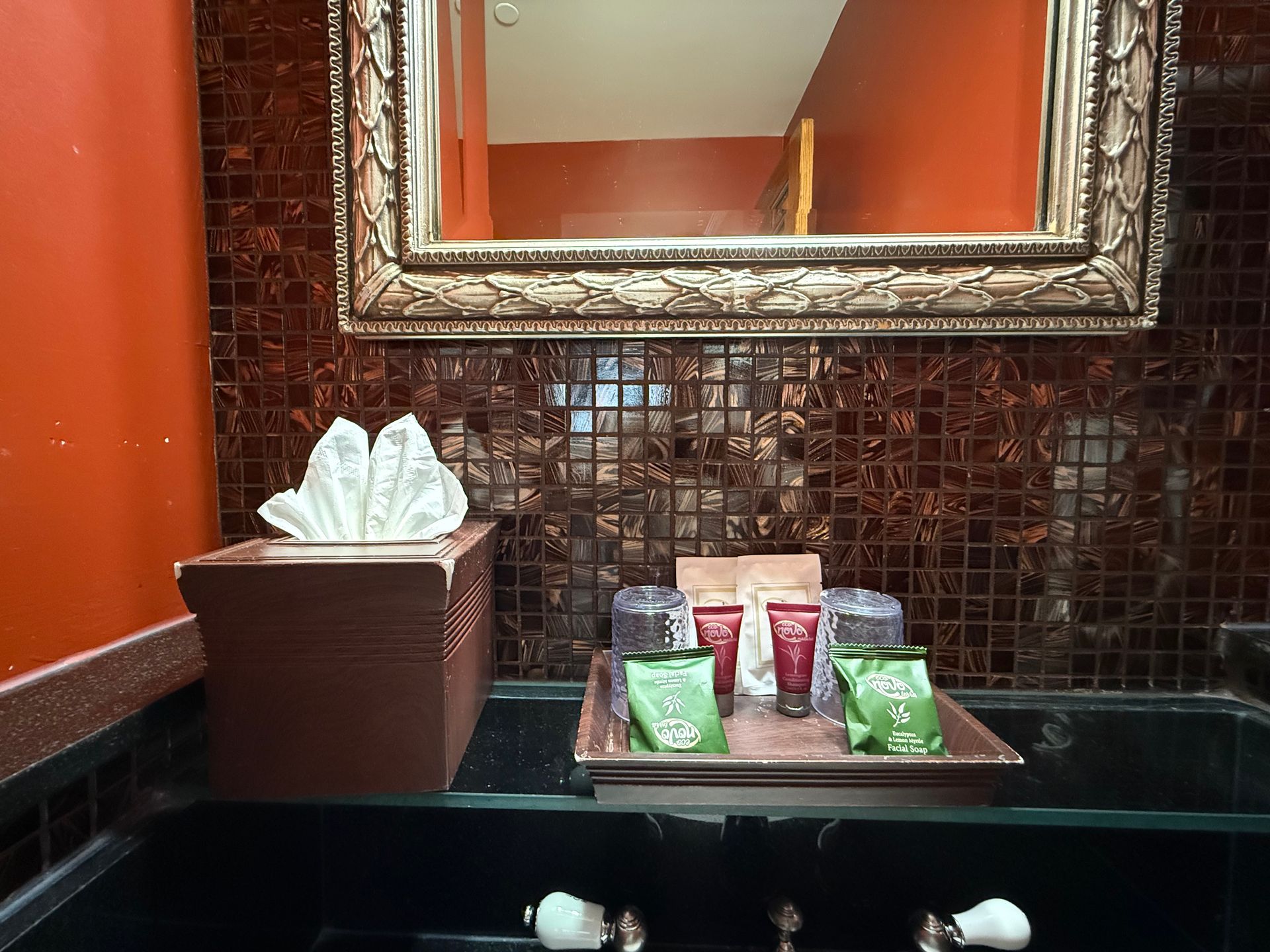 Bathroom vanity with brown tile, mirror, tissues, toiletries, and black countertop.