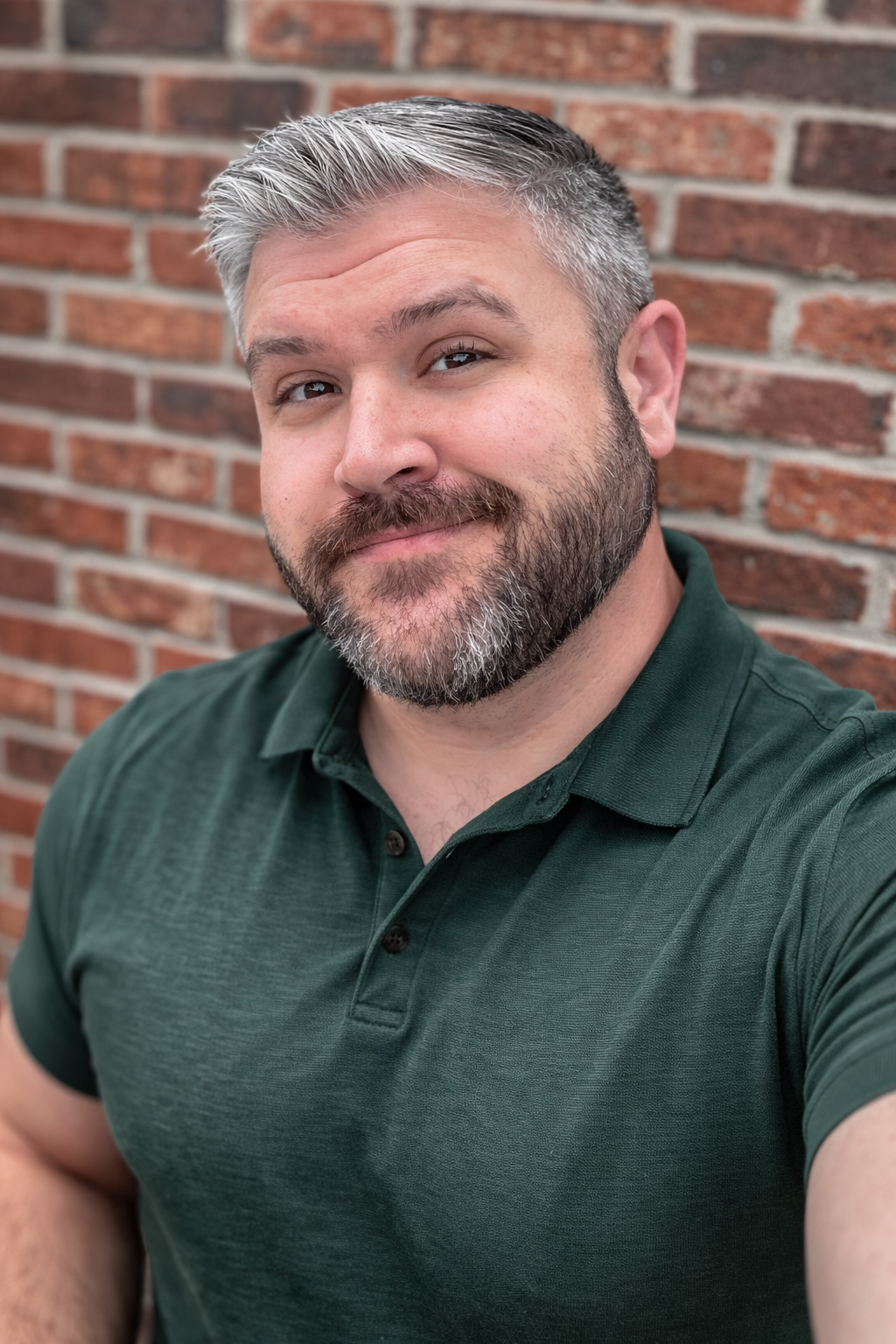 Man in a dark green polo smiling in front of a brick wall