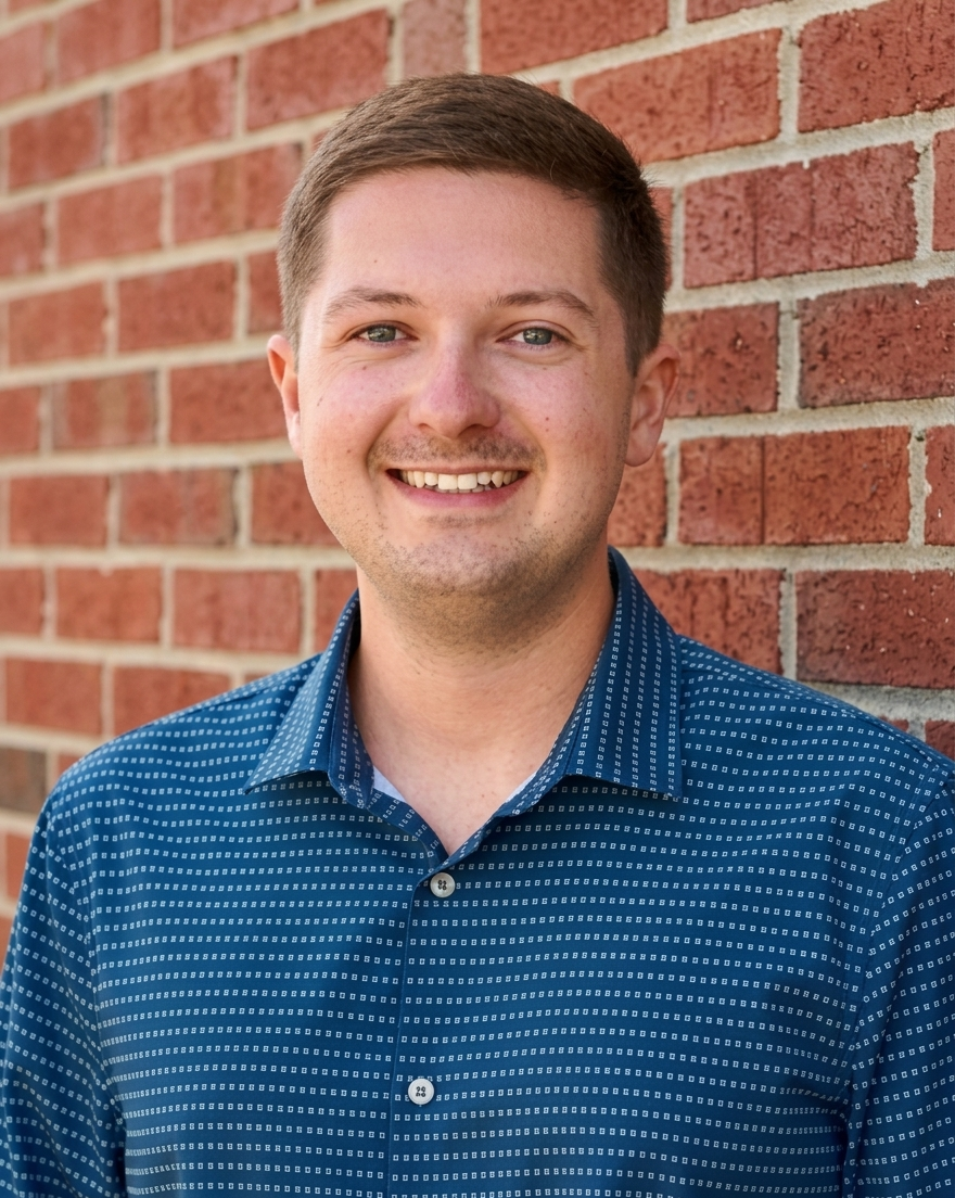 Smiling man in a blue patterned shirt standing against a brick wall