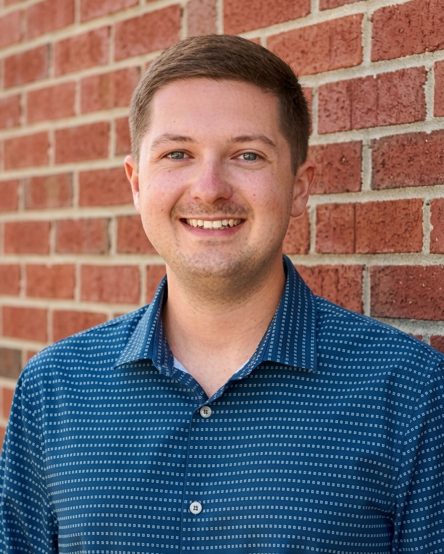 Smiling person in a blue patterned shirt standing against a red brick wall