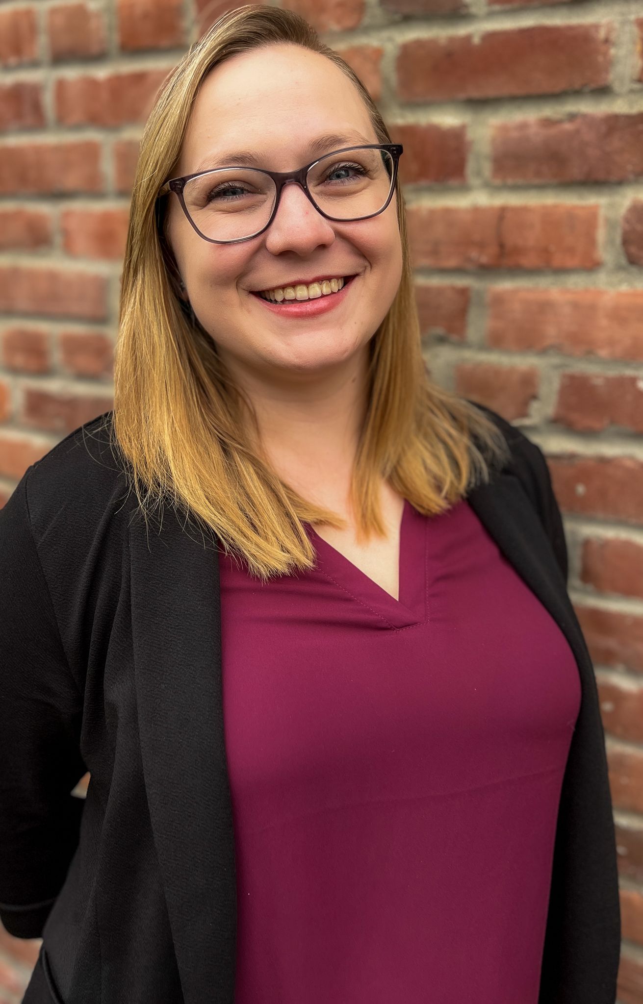 Smiling woman in glasses wearing a maroon top and black cardigan in front of a brick wall