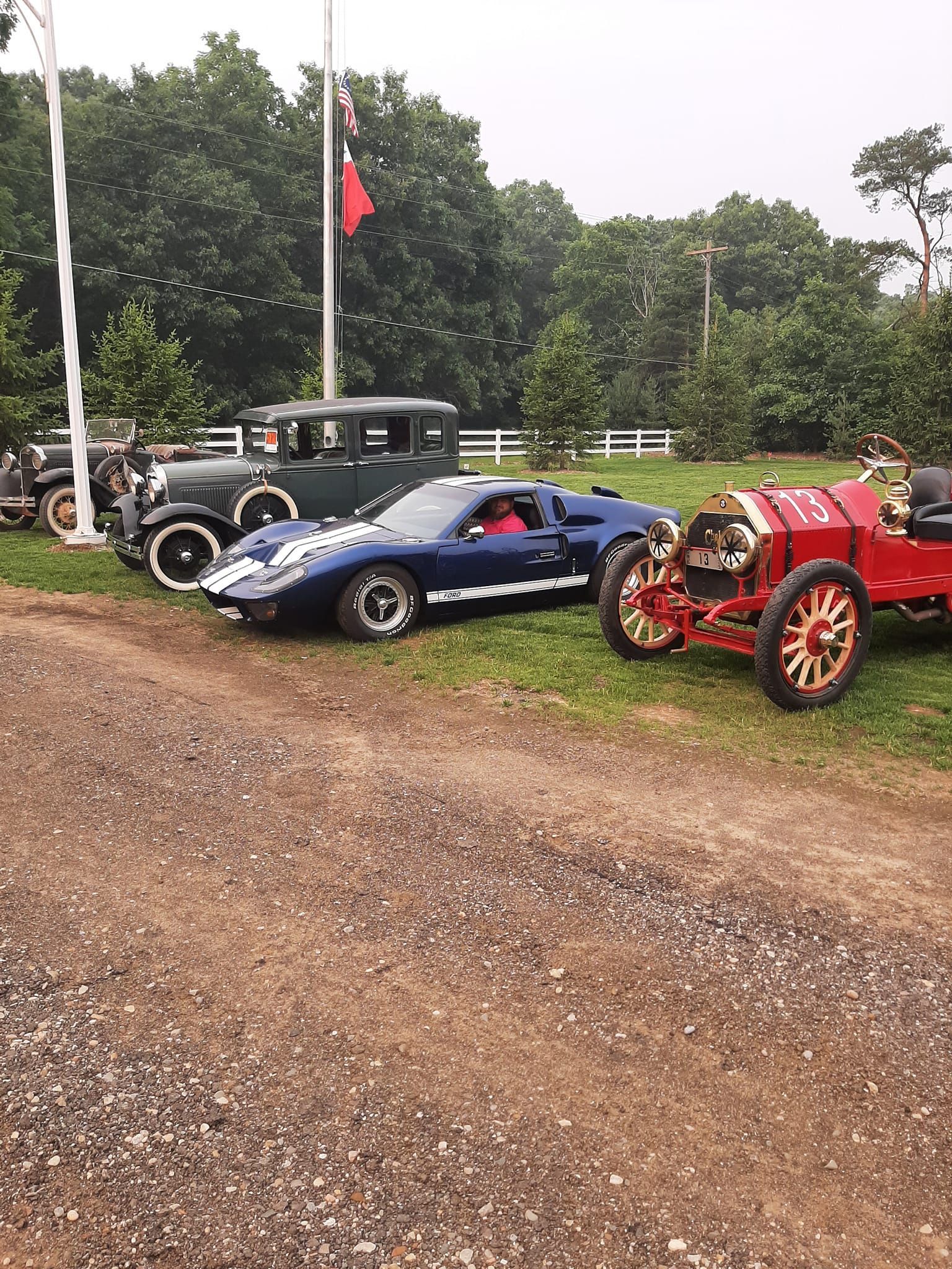 Vintage cars parked on a gravel lot, including a blue coupe and red race car, with trees and a black antique car behind.
