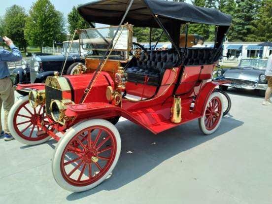 Vintage red open-top car with white wheels and black canopy parked outdoors at a car show