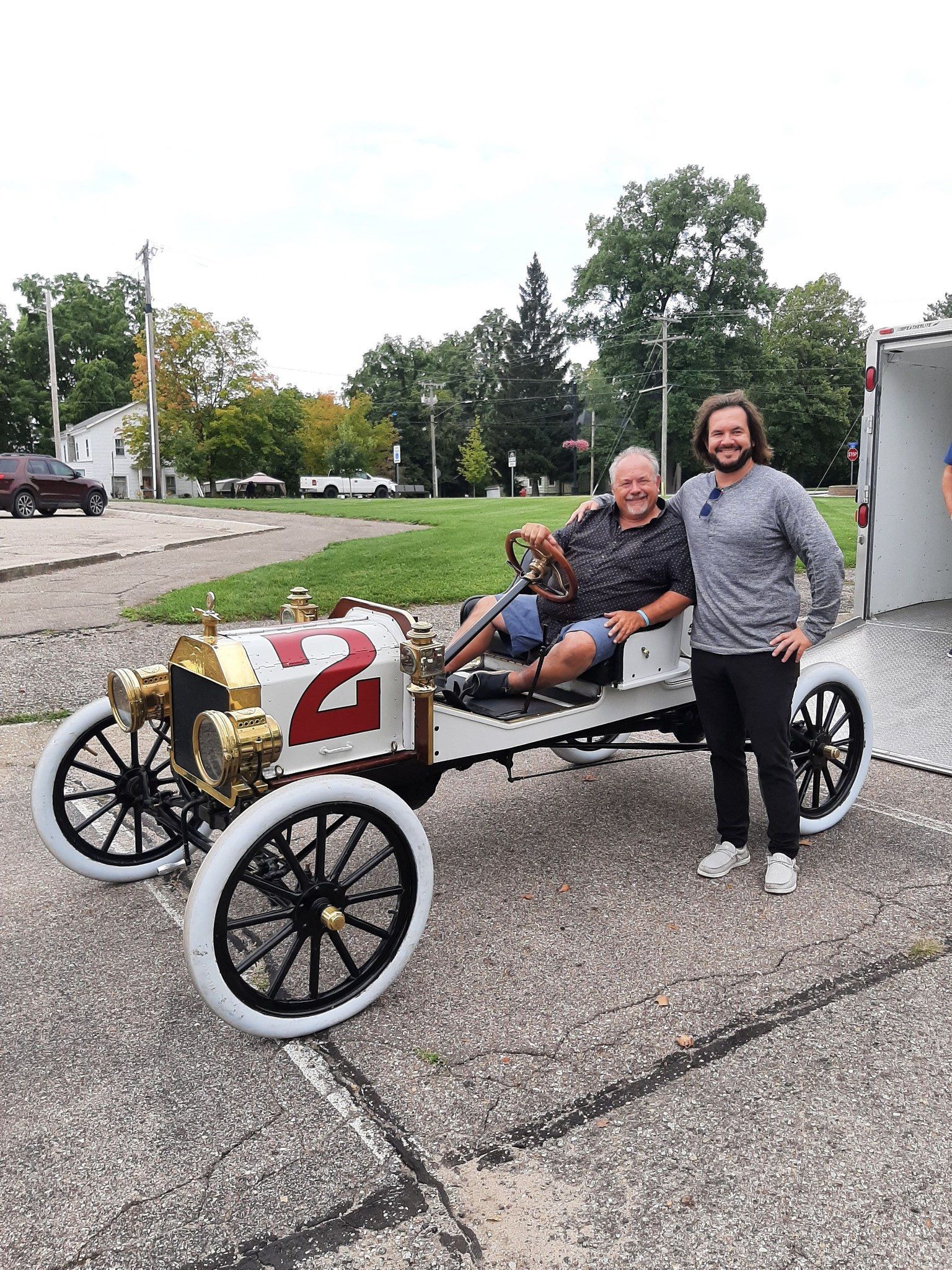 Two people pose beside a vintage white race car with a red number 2 on a paved lot.