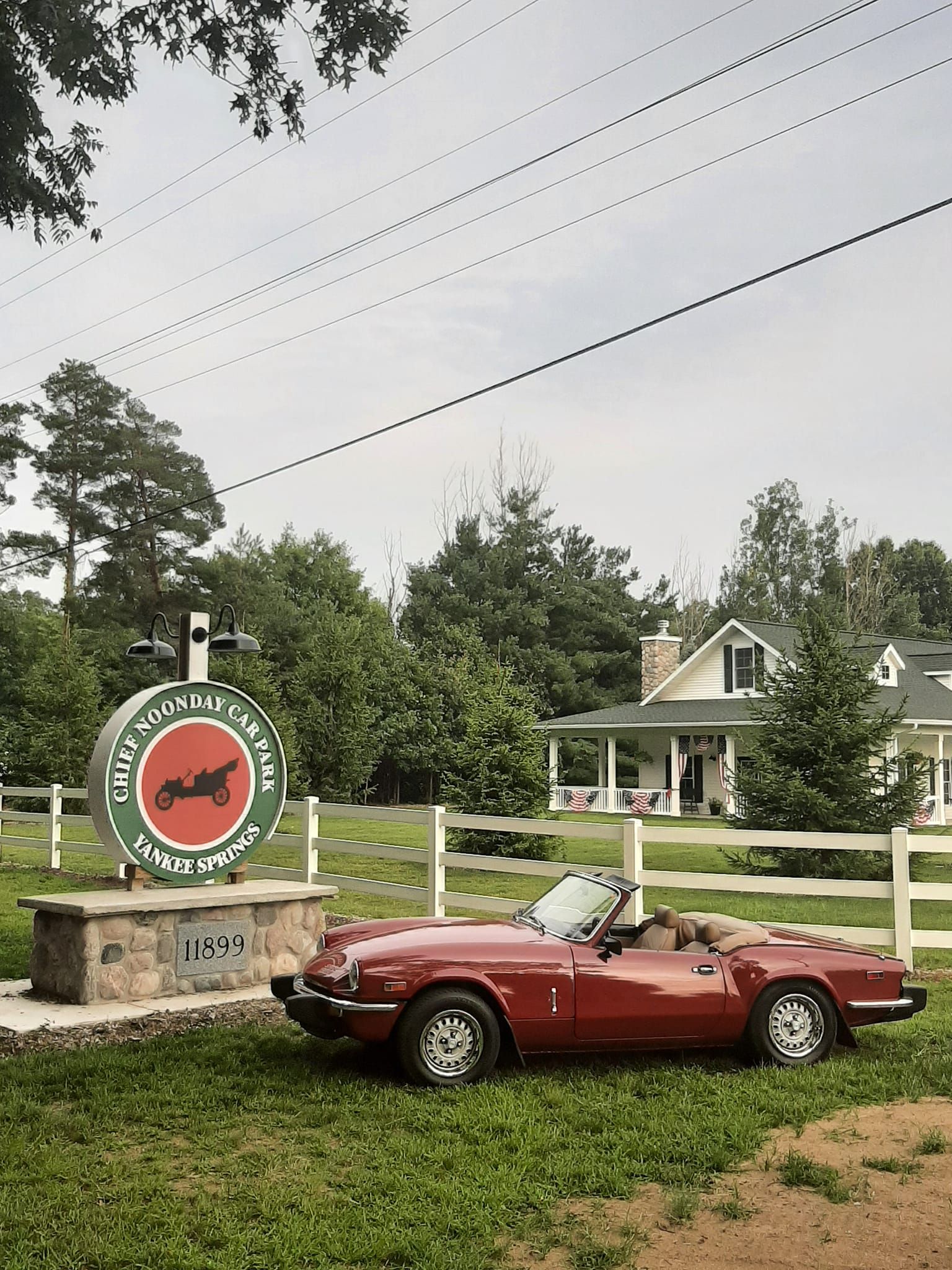 Red vintage convertible parked by a roadside sign and white fence in a wooded neighborhood
