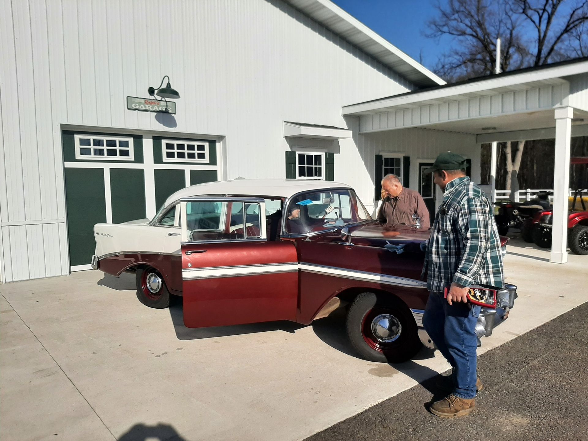 Classic burgundy-and-white vintage car on display outside a white building, with two people inspecting it