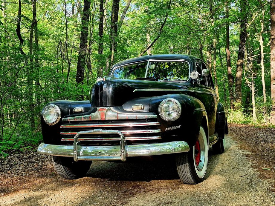 Black vintage pickup truck on a forest dirt road surrounded by green trees.