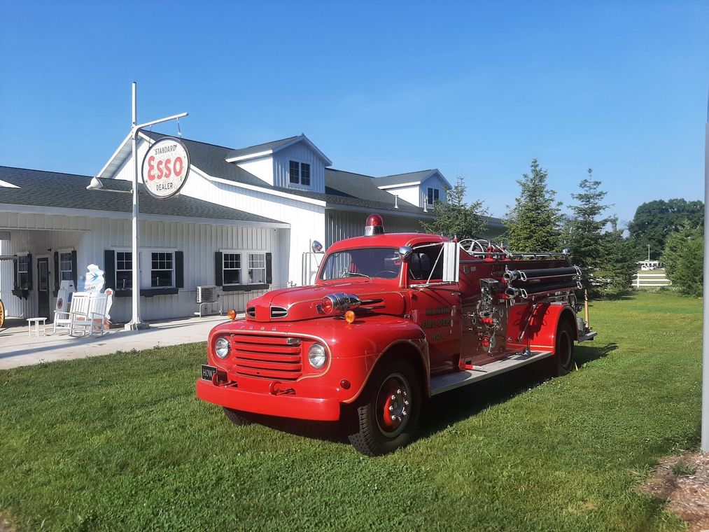 Red vintage fire truck parked on grass beside a white storefront with an ESSO sign