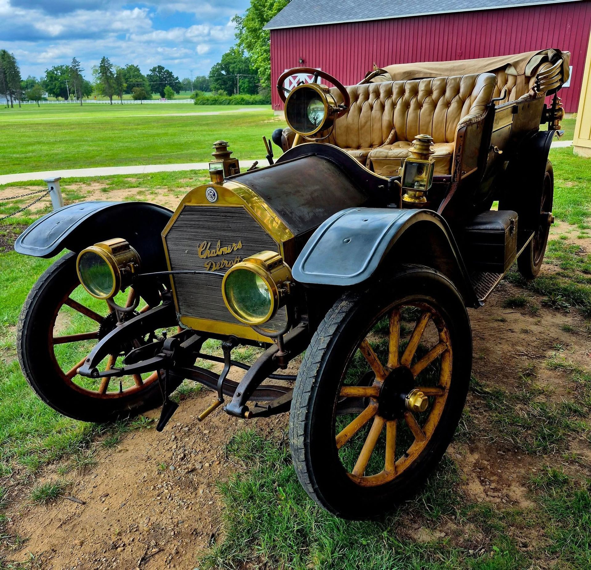 Antique vintage car with tan tufted seats parked on grass beside a red barn under a blue sky