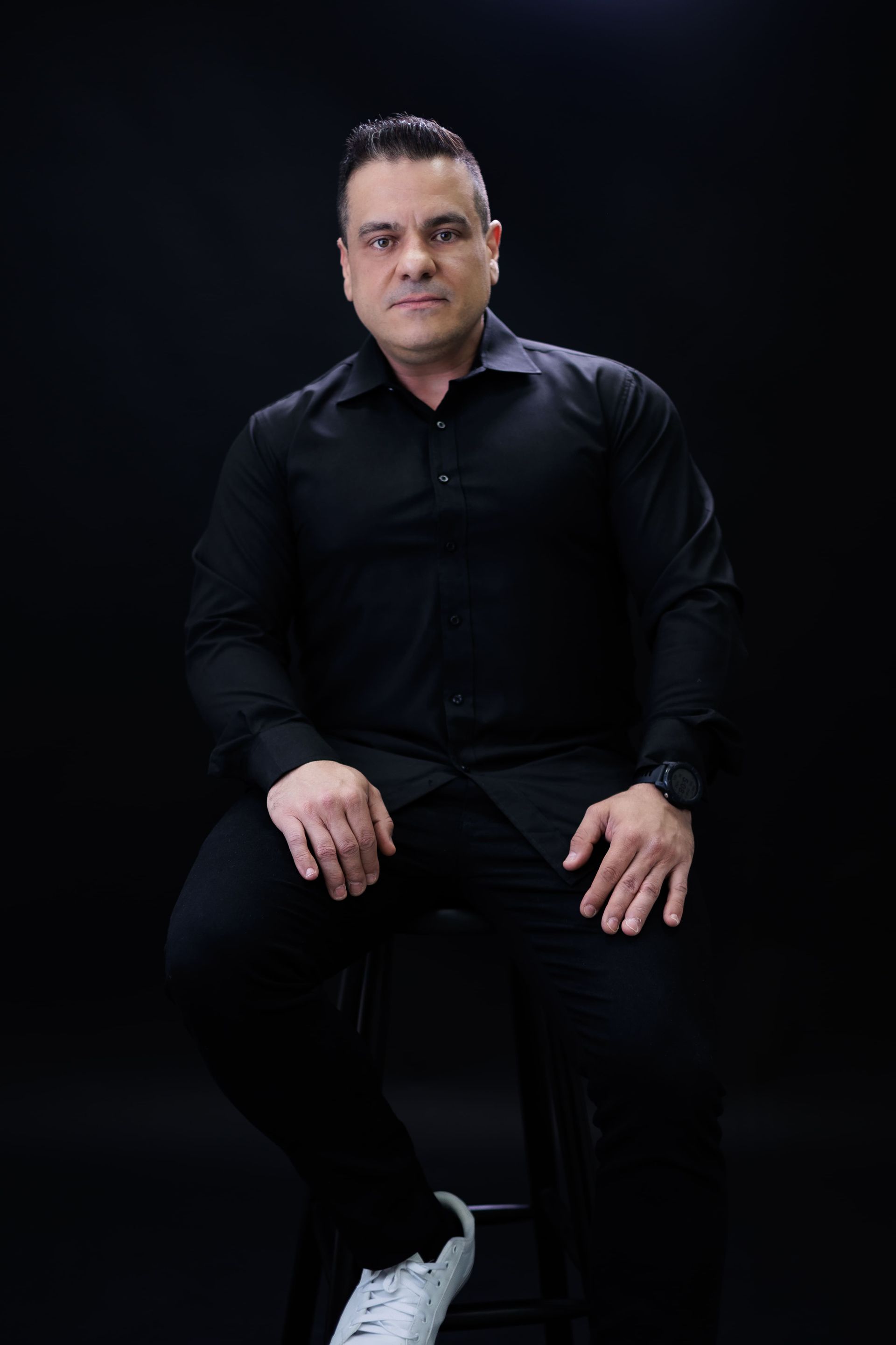 Homem de camisa e calça pretas sentado em um banquinho contra um fundo escuro.