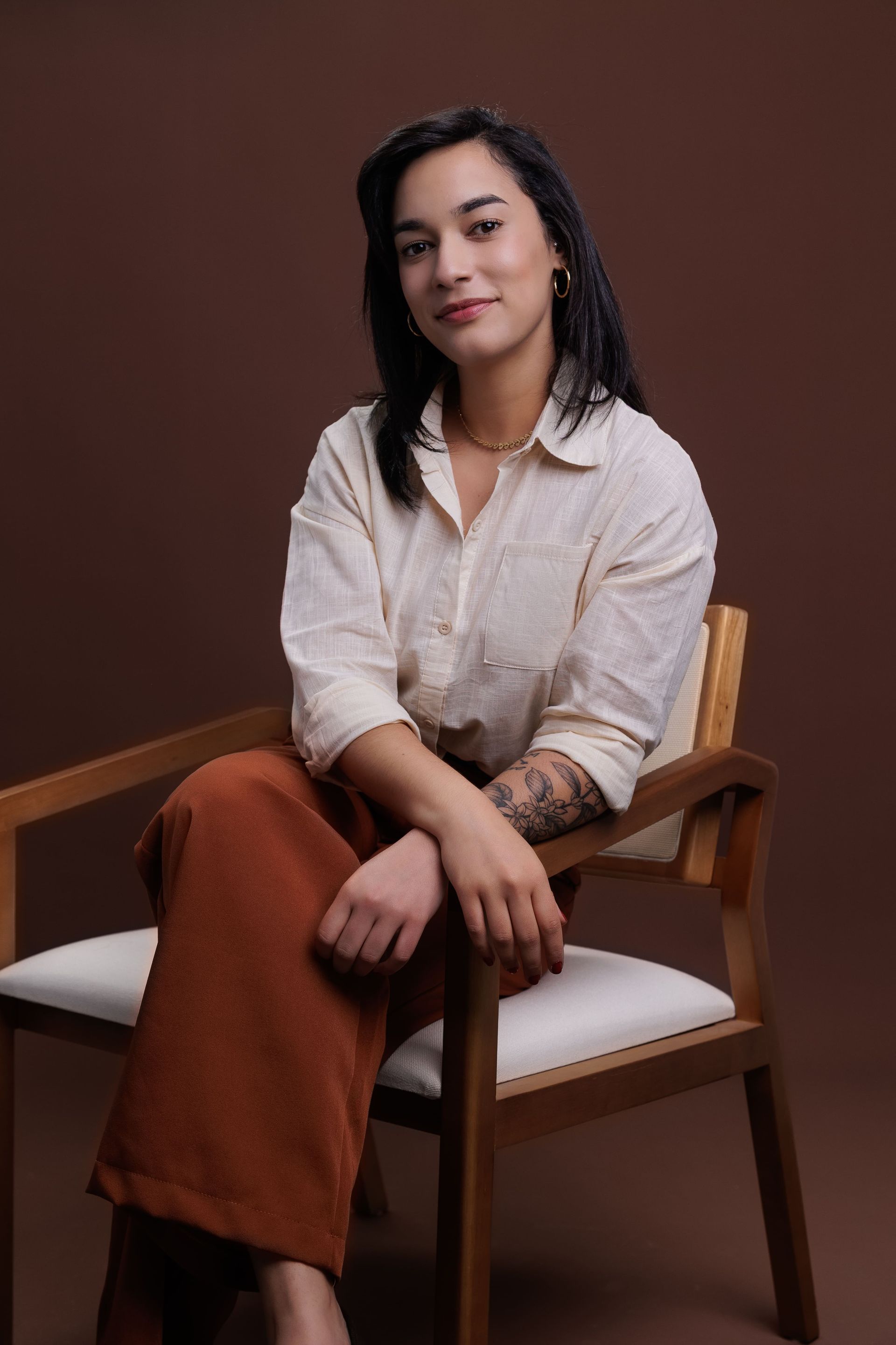 Mulher sentada em uma cadeira, vestindo camisa creme, calças ferrugem, tatuagens nos braços; contra um fundo marrom.