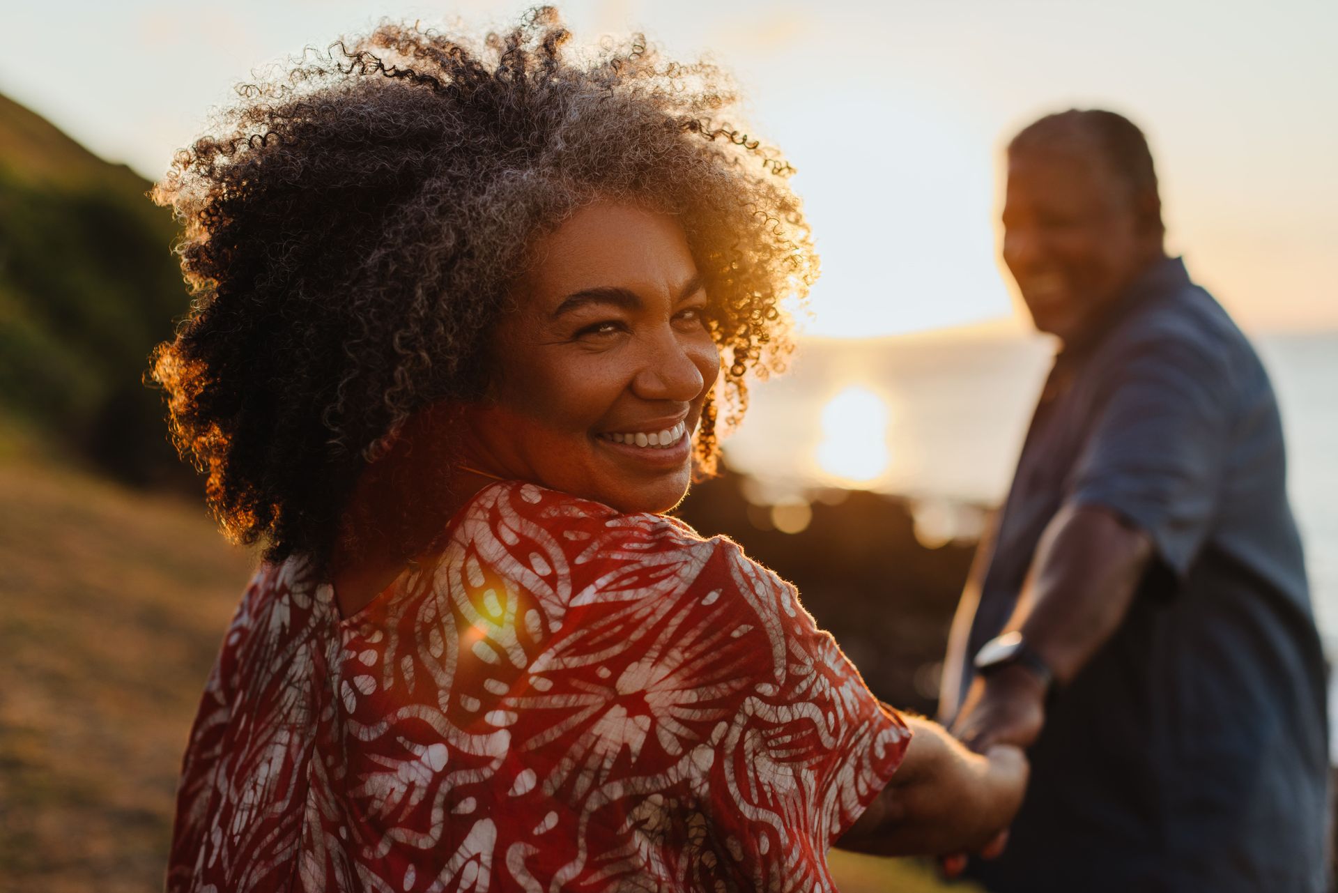 Mulher sorridente, de mãos dadas com um homem desfocado, olha por cima do ombro para o pôr do sol perto da água.