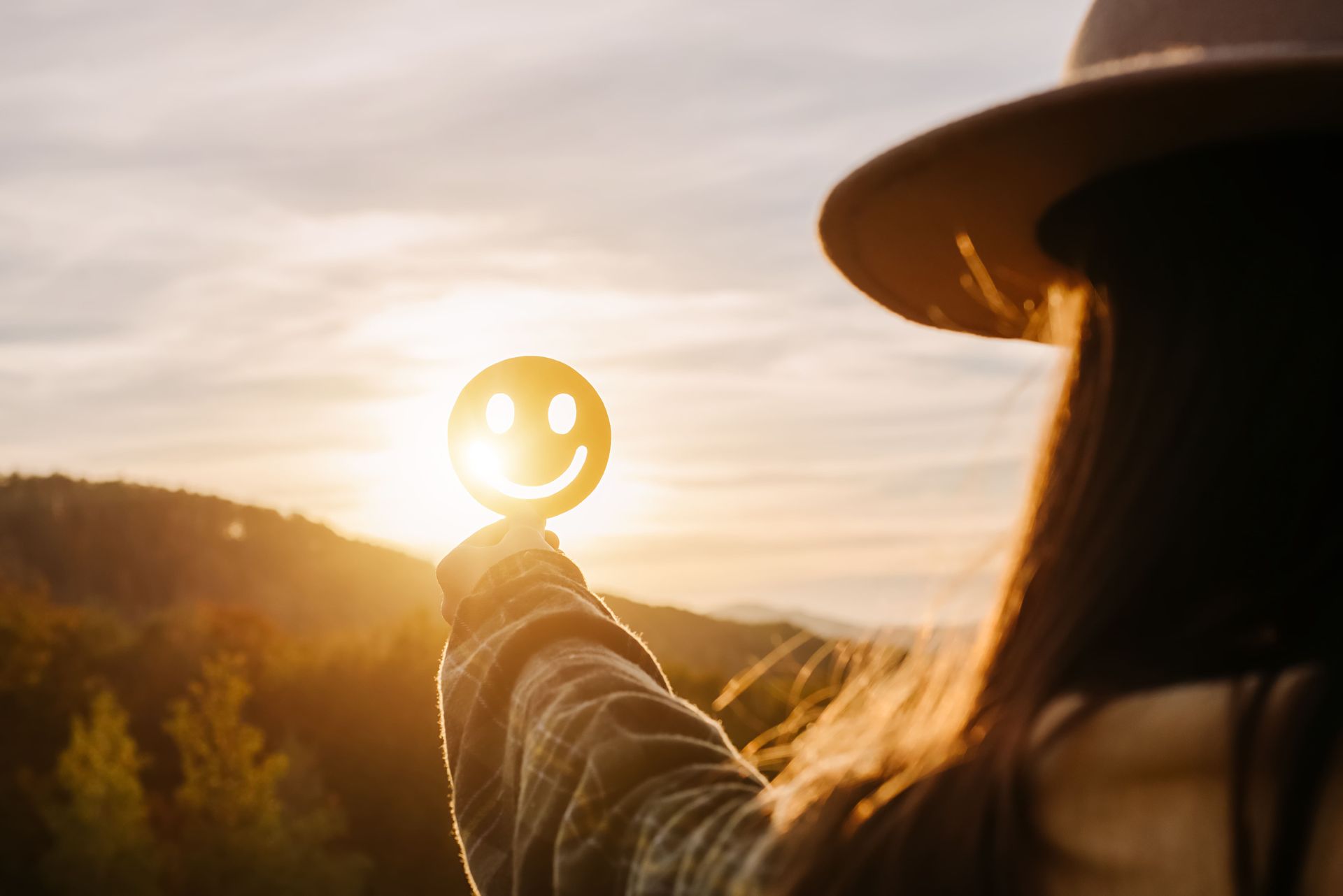 Mulher segurando um recorte de rosto sorridente contra o sol, com vista para a montanha.