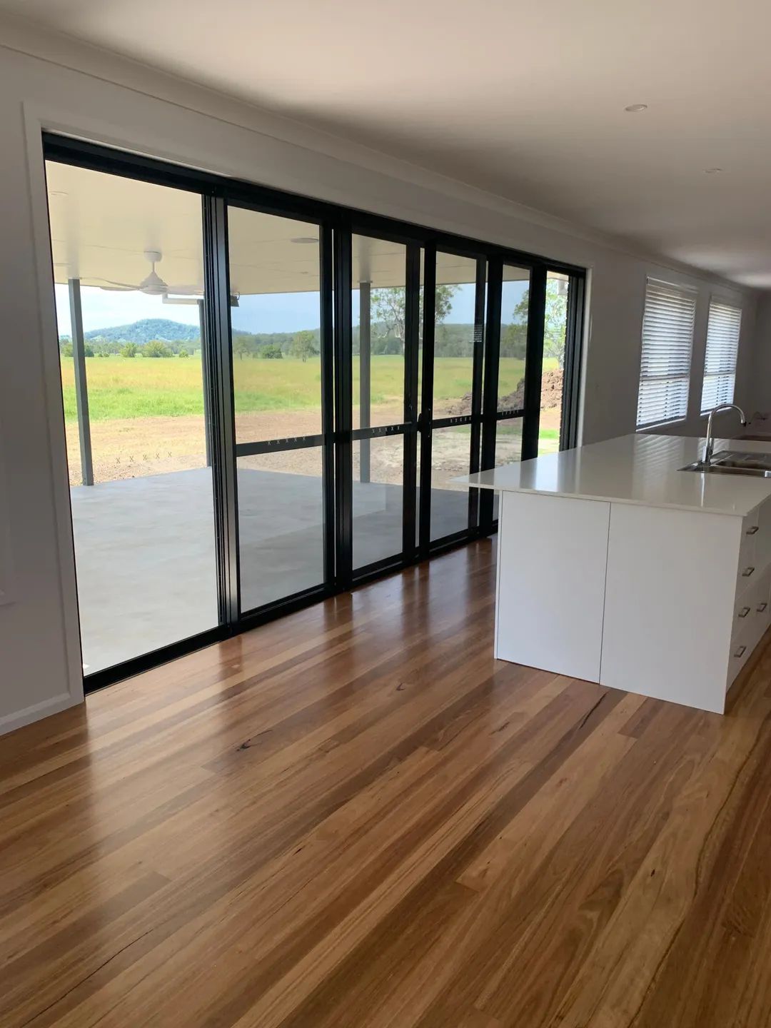 Interior with wood floor, white kitchen island, and large black-framed glass doors opening to a patio — C & S Building In Bentley, NSW