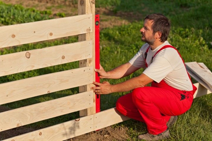 Man in red overalls using a level on a wooden fence outdoors.