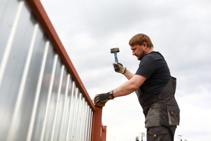 Man in work clothes hammering on a red metal structure, outdoors against a cloudy sky.