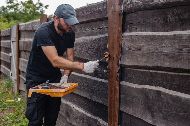 Man in black clothing painting a wooden fence brown outdoors.