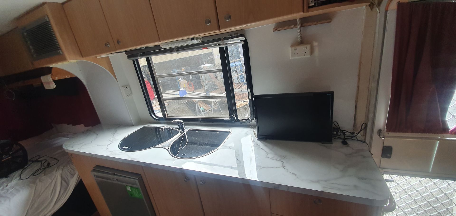 Interior of a camper, showing a countertop with a sink, window, and small TV. Beige cabinets and a curtain are visible.