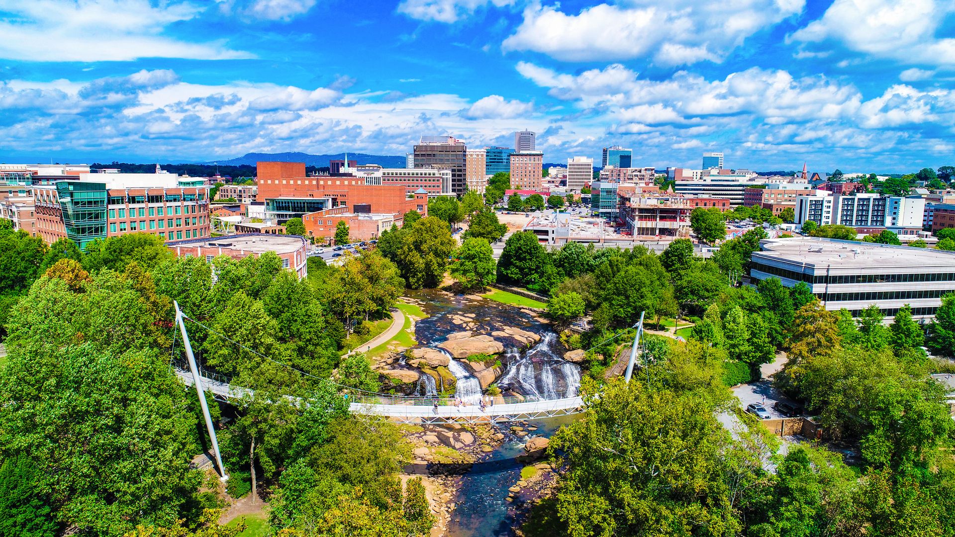 City skyline beyond a lush green park with a waterfall and a river under a bright blue sky