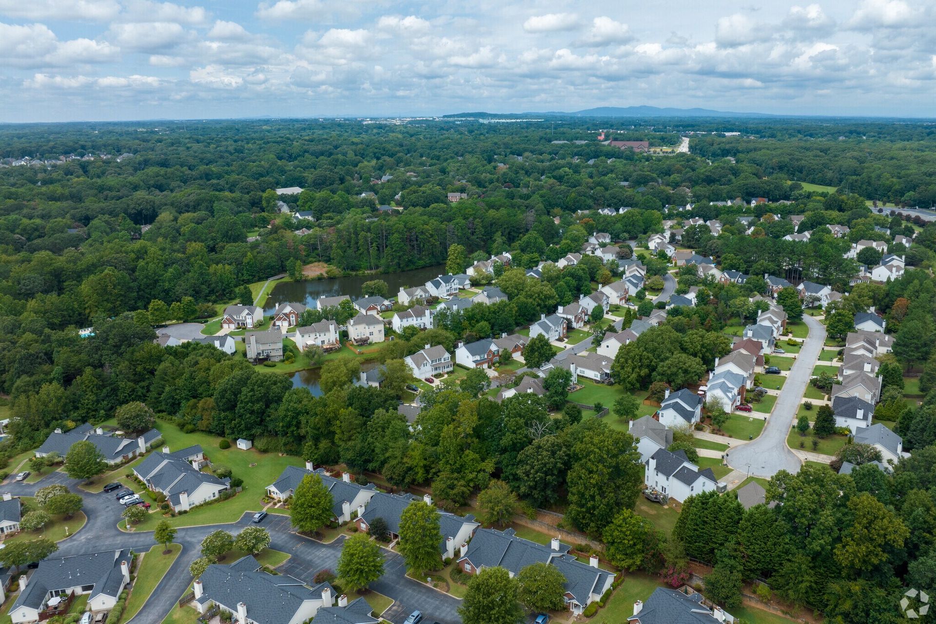 Aerial view of a suburban neighborhood surrounded by dense green trees under a cloudy blue sky