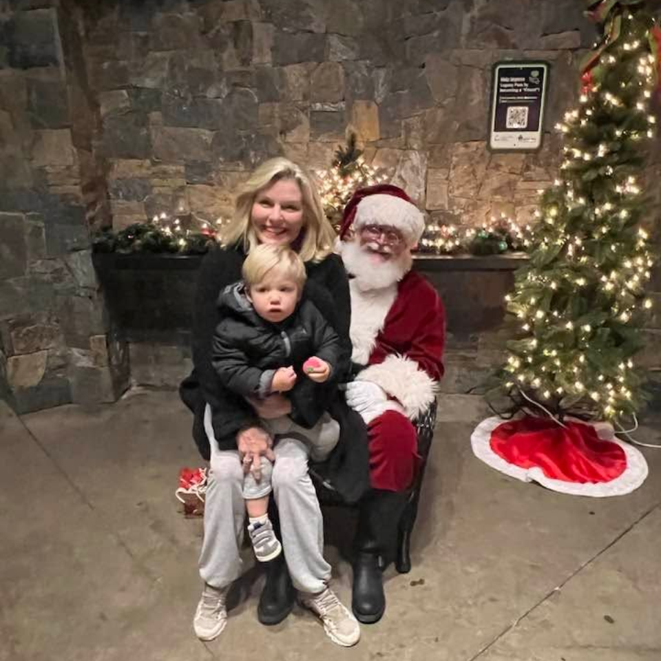 Woman and child posing with Santa beside a lit Christmas tree and stone wall