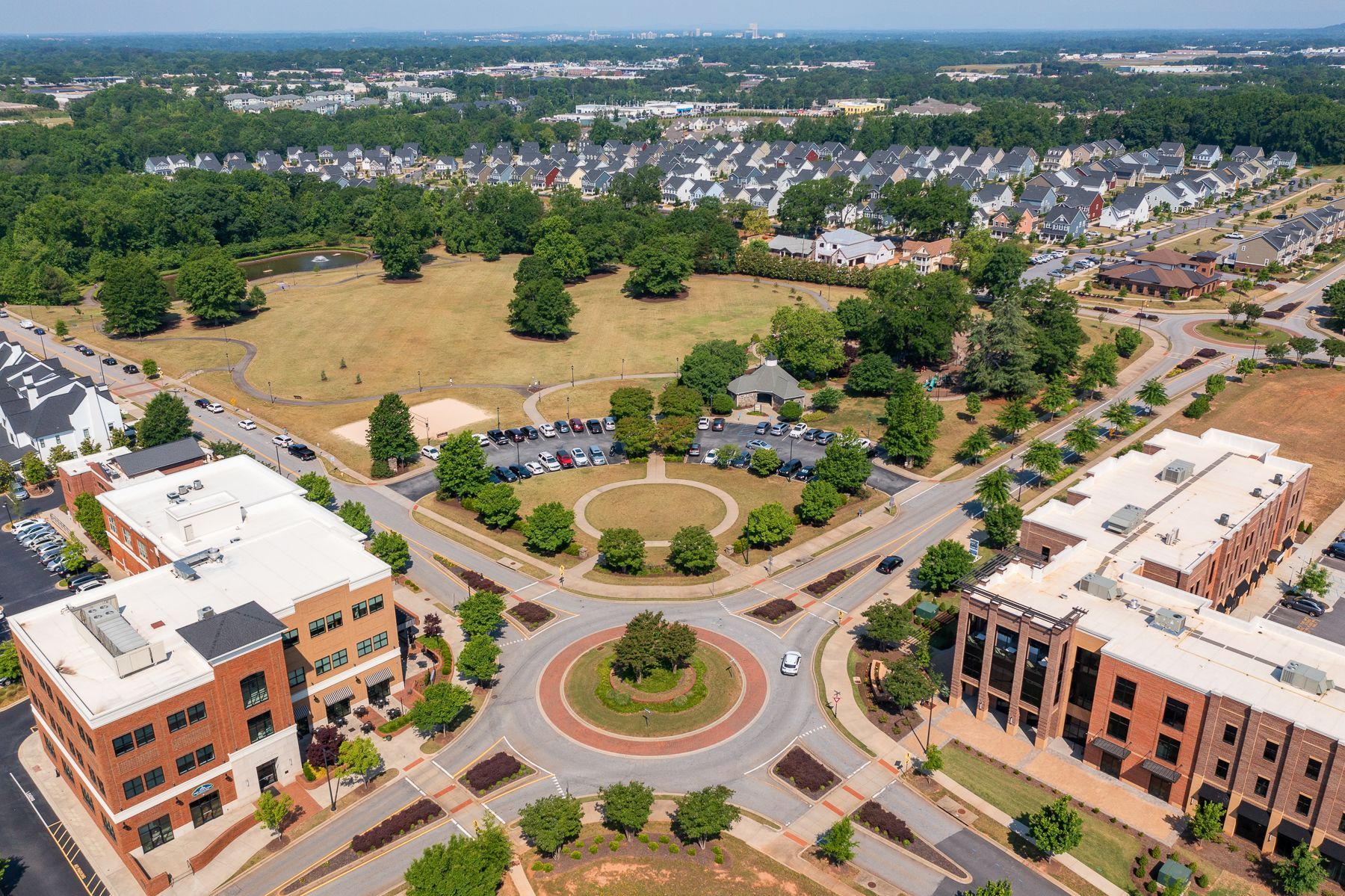 Aerial view of a suburban roundabout with buildings, trees, and parking lots under a clear sky