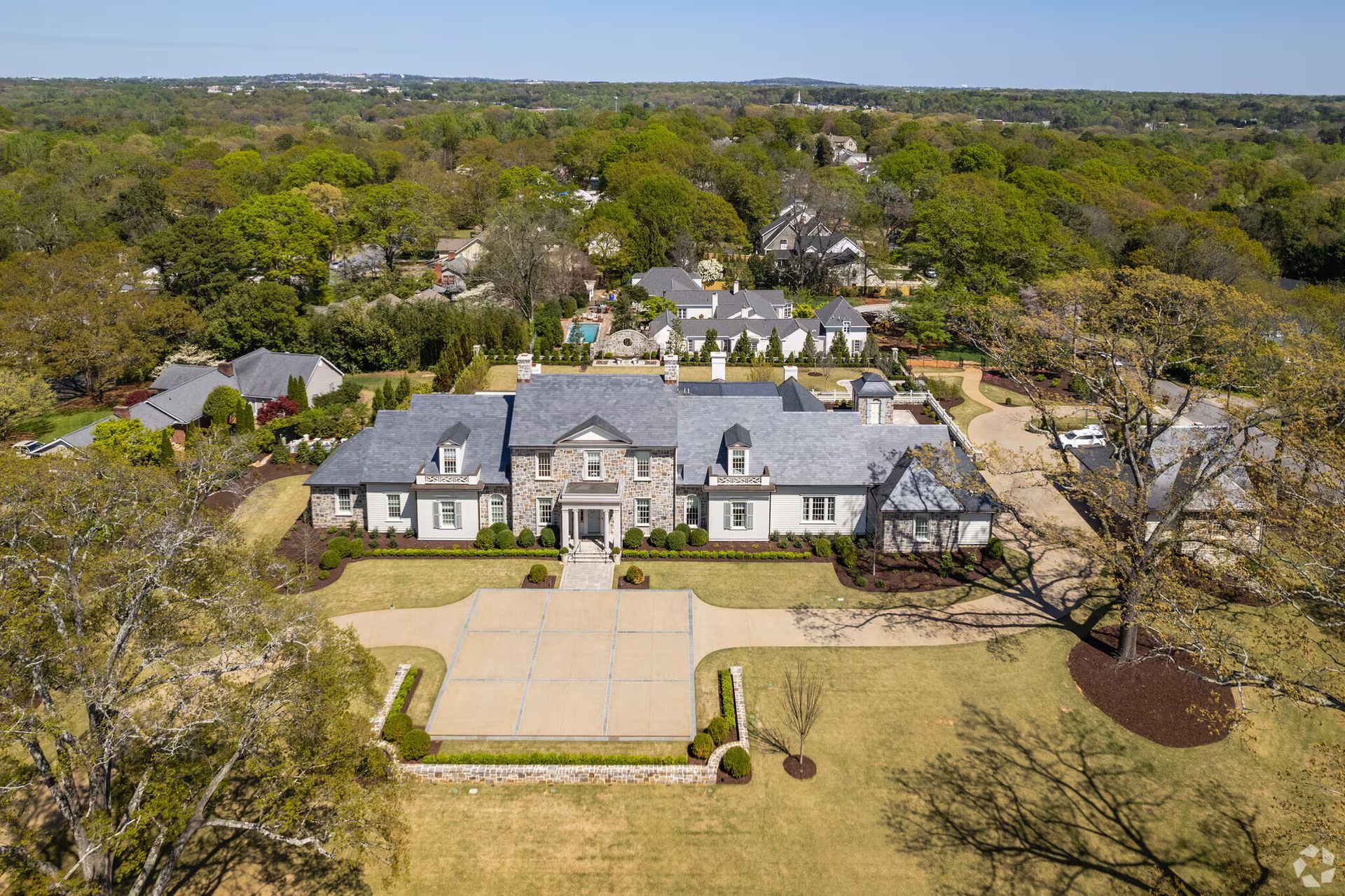 Aerial view of a large gray mansion with a circular driveway, surrounded by trees and open land