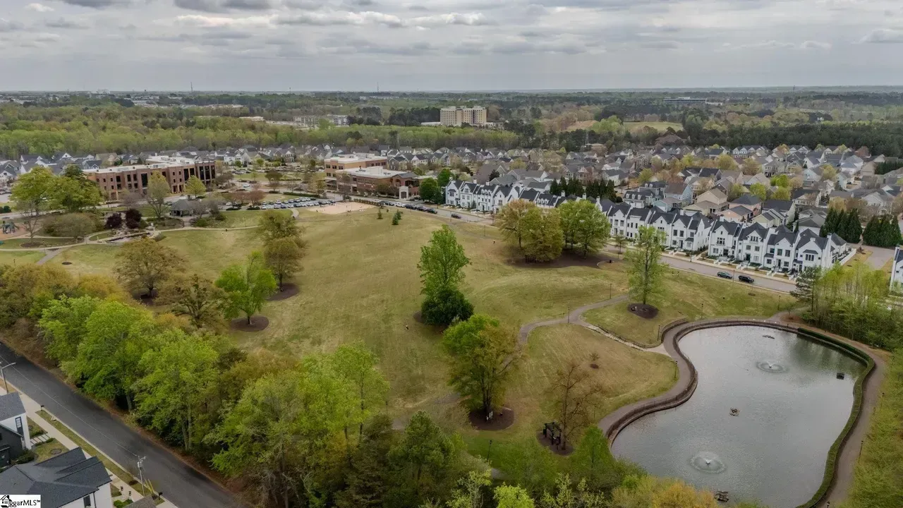 Aerial view of a park with open grass, trees, a pond, and houses in the background.