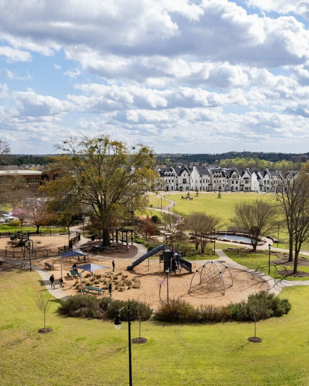 Park playground with slides and climbing structures in a grassy field, with houses and cloudy sky beyond.