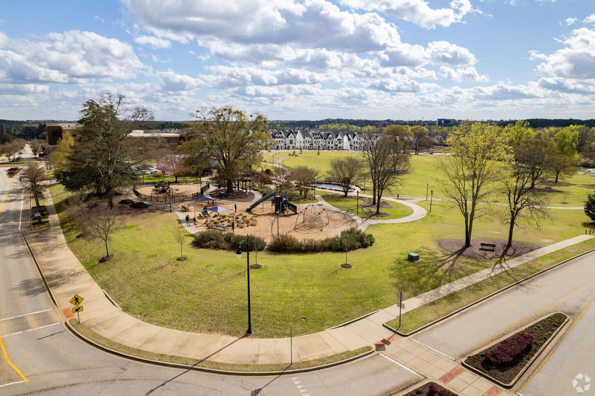 Park with a walking path, trees, and a central sandy play area under a cloudy sky