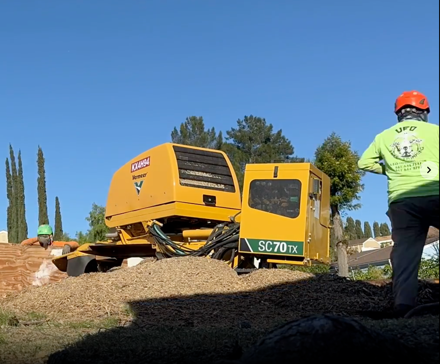 Yellow tracked construction machine on a dirt mound, with a worker in a neon shirt and orange helmet nearby.