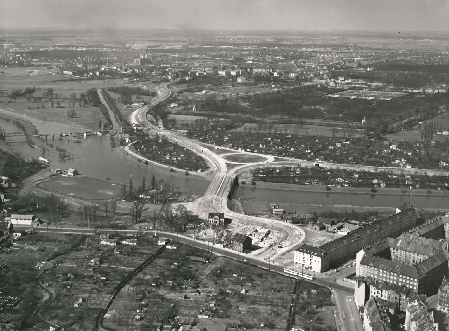 Freigegeben durch das Historische Museum Hannover. Luftbild von Luftreisedienst Niedersachsen GmbH., Hann., Frühjahr 1958: Blick auf die Leine mit Schwanenburgbrücke Westschnellweg kurz vor der Eröffnung am 1.4.1958, Schwanenburgkreisel und die Stadtteile Limmer (Leine-Verbindungs-Kanal und Leinewehr), Linden-Nord (Limmerstraße, Steigerthalstraße), Herrenhausen (Großer Garten Herrenhausen, Heizkraftwerk, Schleuse zum Ernst-August-Kanal) und Nordstadt (Wasserkunst, Westschnellweg, Schwanenburgkreisel, Bremer Damm). Ein Haus auf der Trasse hindert noch den Weiterbau der Westtangente (Westschnellweg) nach Süden zum Deisterplatz. Auf der Höhe der Straßenbahn im Vordergrund wird drei Jahre später auf dem Kleingartengelände das Freizeitheim Linden eröffnet. Bildarchiv HMH. Scan vom Foto 21×29 cm. Freigegeben durch das Historische Museum Hannover.