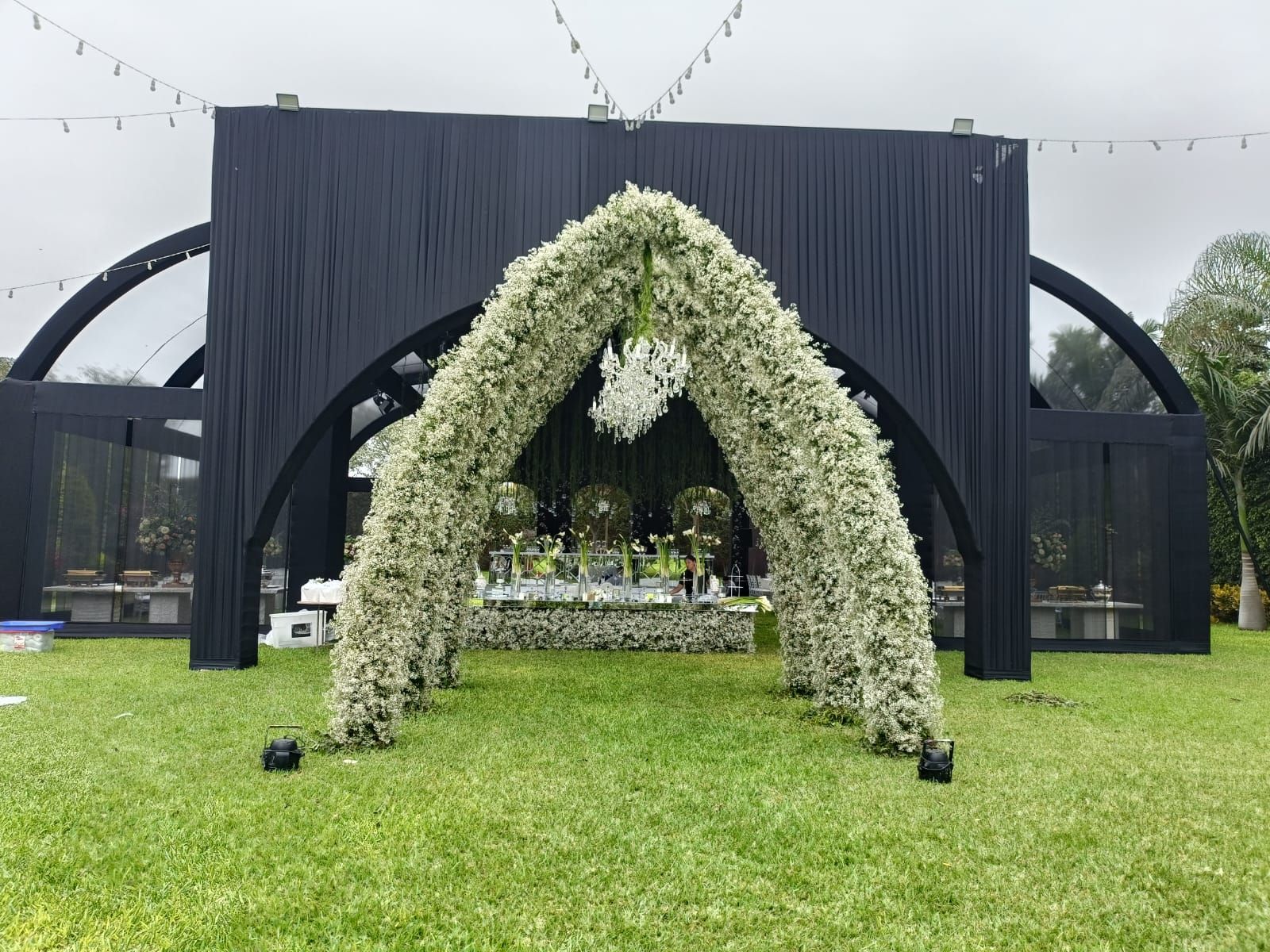 Arco floral blanco sobre un césped verde frente a una carpa negra para eventos al aire libre.