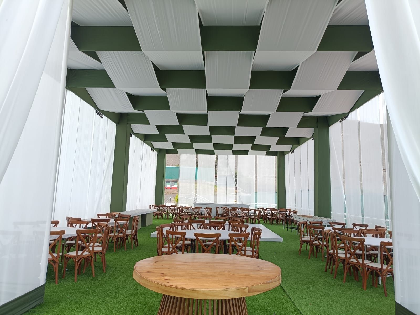 Outdoor event space with checkered green-and-white canopy, wooden tables, and chairs on grass.