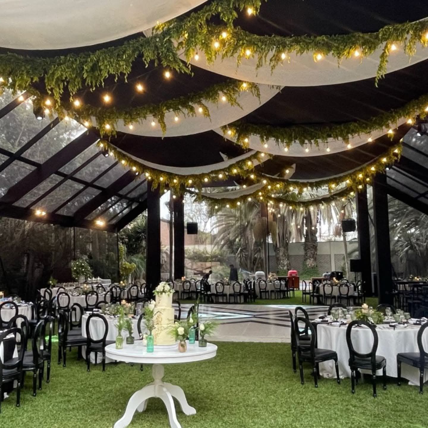 Espacio para eventos al aire libre decorado con guirnaldas de luces, cortinas blancas, sillas negras y mesas sobre césped.