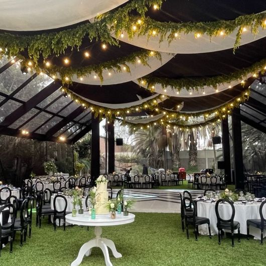 Elegante carpa para eventos al aire libre con guirnaldas de luces, cortinas blancas y mesas redondas sobre un césped.