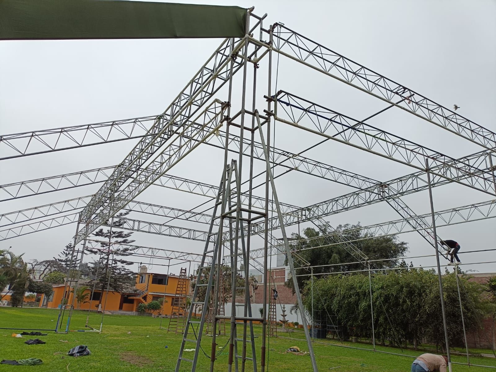 Estructura metálica en proceso de montaje sobre un campo de hierba, con trabajadores y maquinaria de color naranja al fondo.