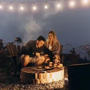 Couple seated by a fire pit on a deck under string lights at dusk, roasting food outdoors.