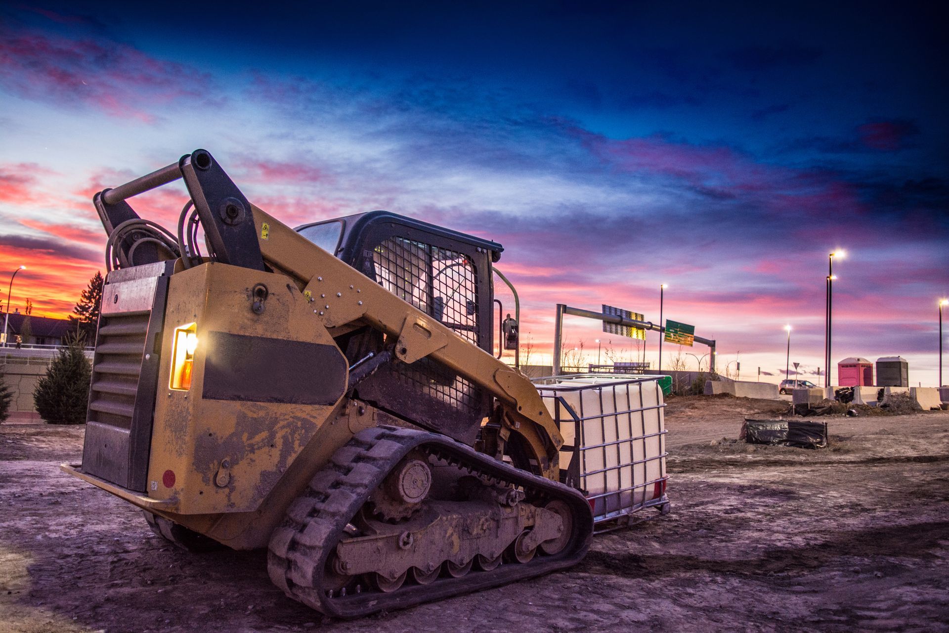 Tracked skid steer loader on a construction site at dusk with colorful sky and street lights.