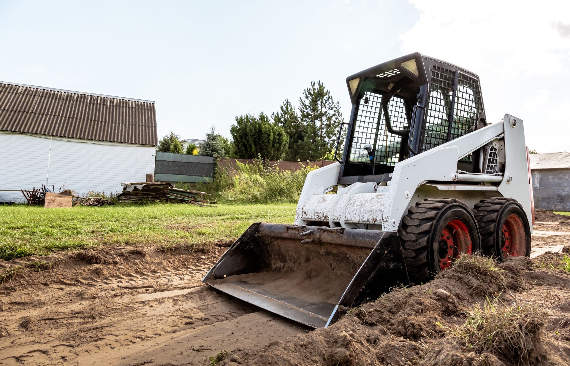 Bobcat skid-steer loader scooping dirt in a muddy field, with buildings and trees in the background.