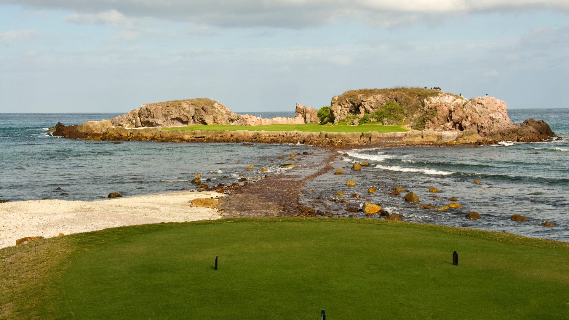 Rocky island in the sea beside a green grassy foreground under a clear sky.