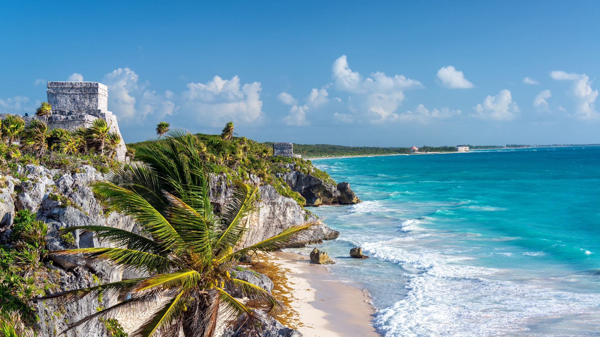 Cliffside ruins above a turquoise beach and palm trees under a bright blue sky
