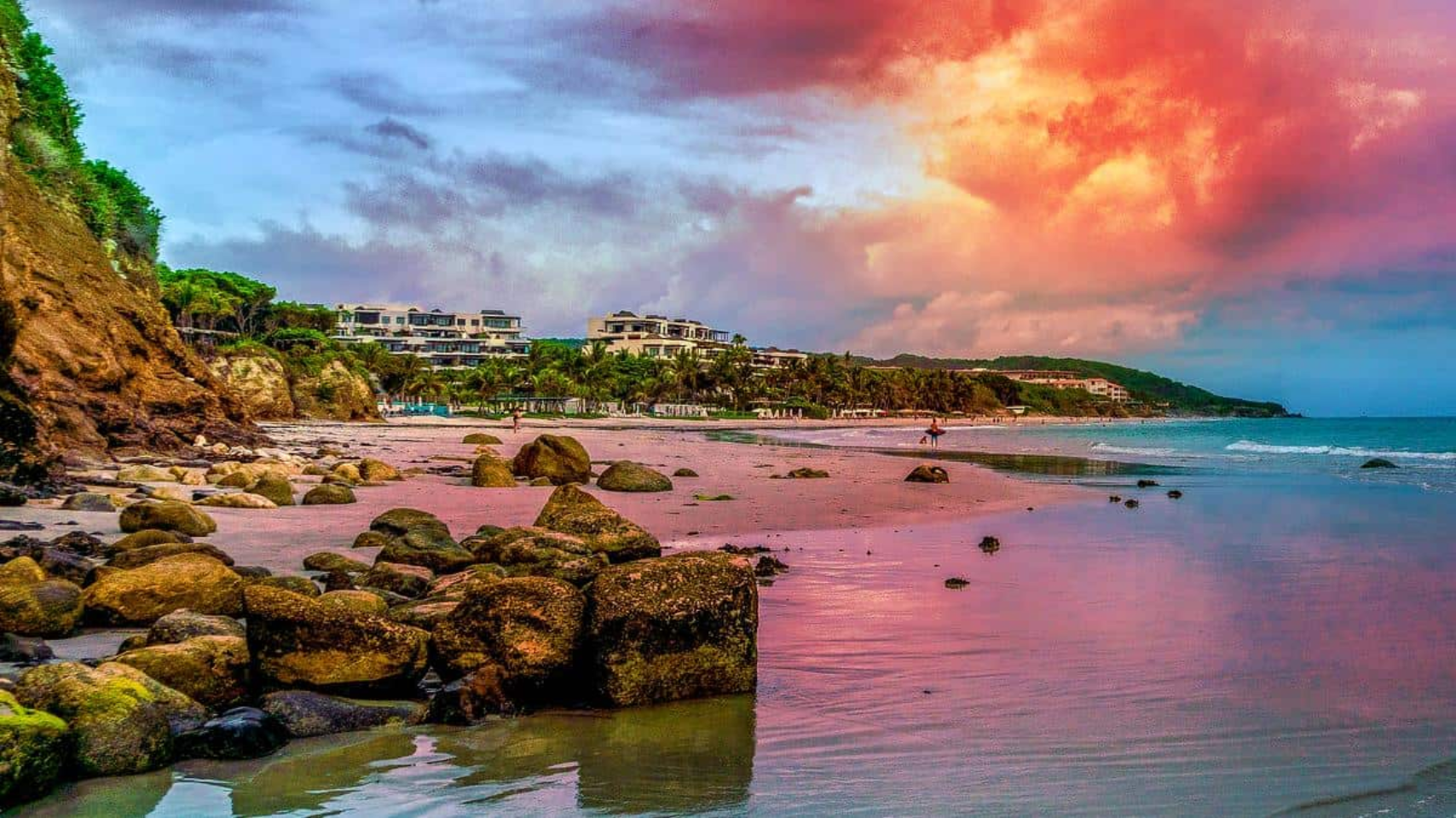 A sunset over a rocky beach with buildings in the background.