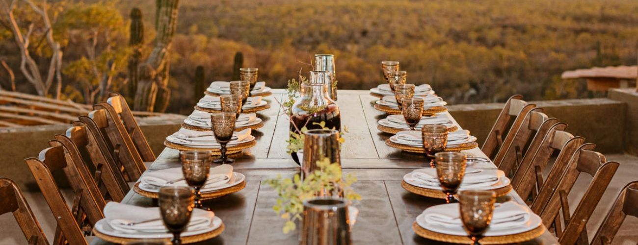 A long wooden table with plates , glasses , and a vase of flowers on it.