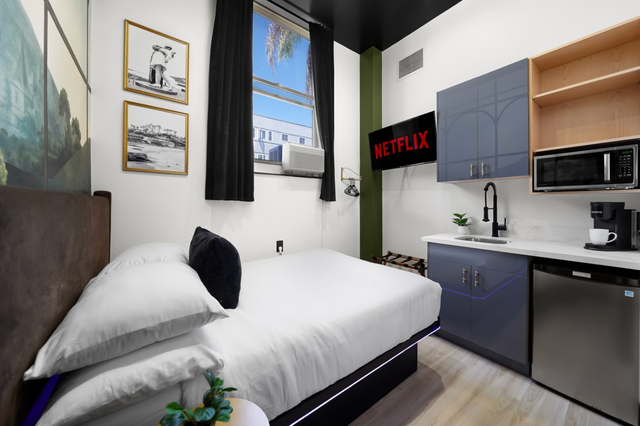 Compact studio room with bed, kitchenette, wall art, and a window overlooking the city.