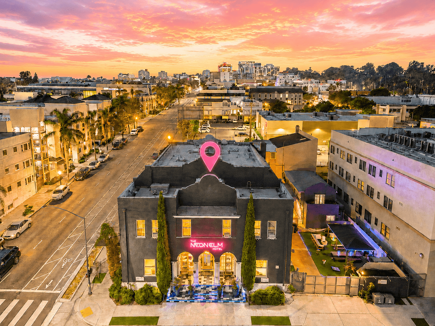 Aerial view of a lit building at sunset with a pink location pin over a city street and skyline.
