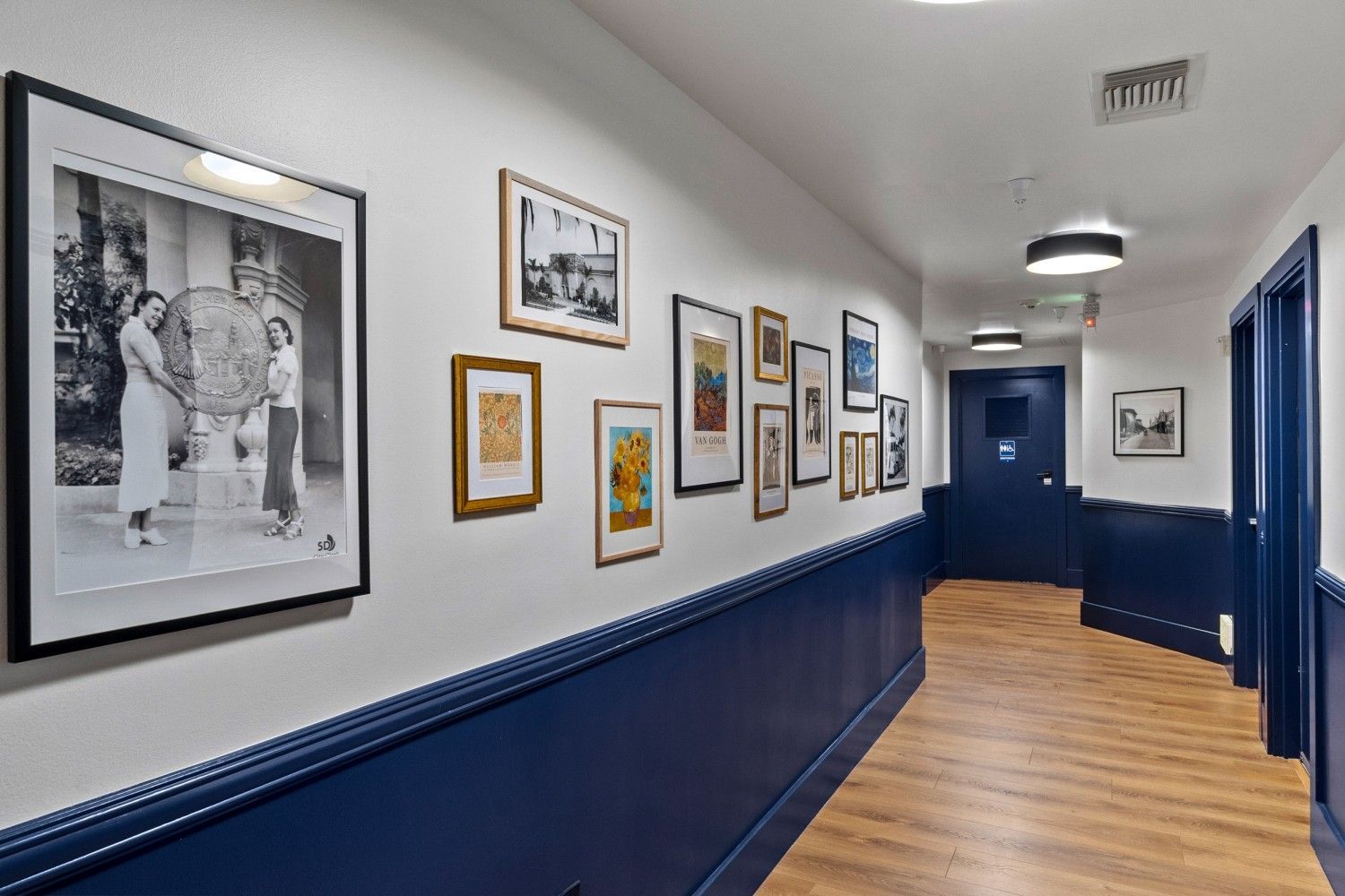 Narrow hallway with framed photos and art, white upper walls, navy lower walls, and wood flooring.